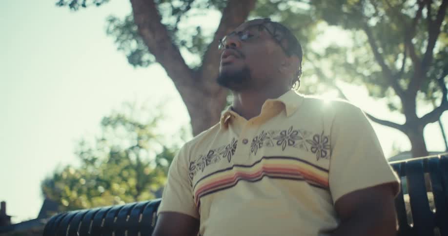 A man with braided hair and glasses wearing a yellow patterned polo shirt sitting on a park bench he is looking at his smartphone with a puzzled or concerned expression gesturing with his hands as if in disbelief or frustration