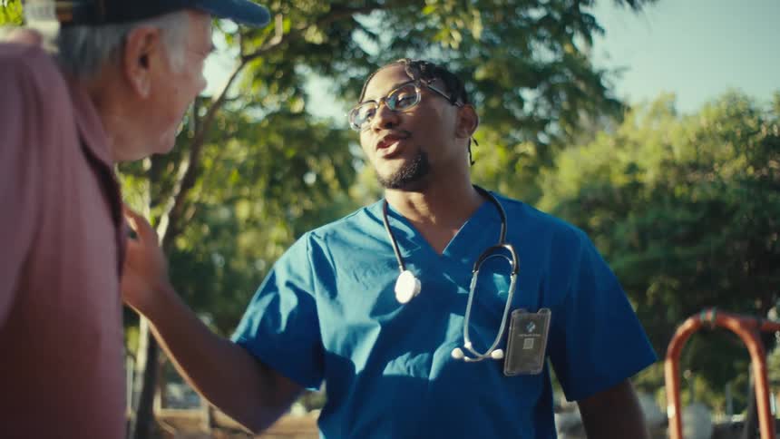 A young man in blue medical scrubs and a stethoscope around his neck is seen talking and smiling with an older man outdoors the scene appears to be set in a sunny park or residential area with trees in the background
