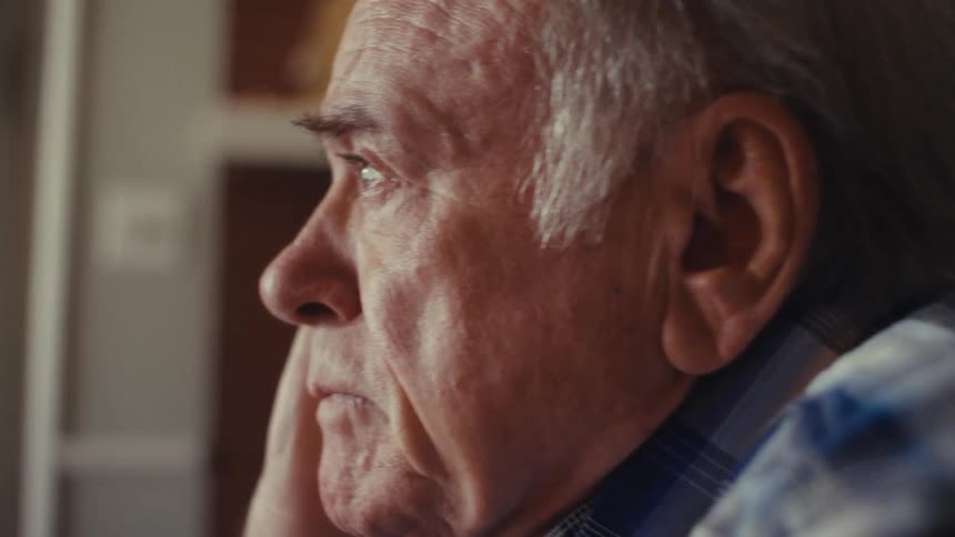 An elderly person in a plaid shirt and white earbuds sits indoors visibly distressed and crying