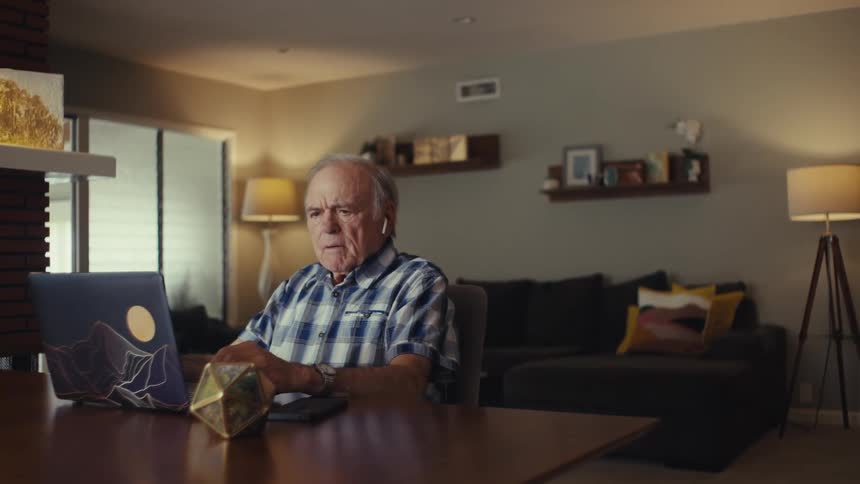 Elderly man wears airpods to take a call while sitting at a table in a living room during daytime
