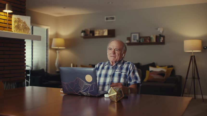 An elderly man in a plaid shirt laughs with both arms raised wearing earbuds and seated at a table on a video call with a laptop