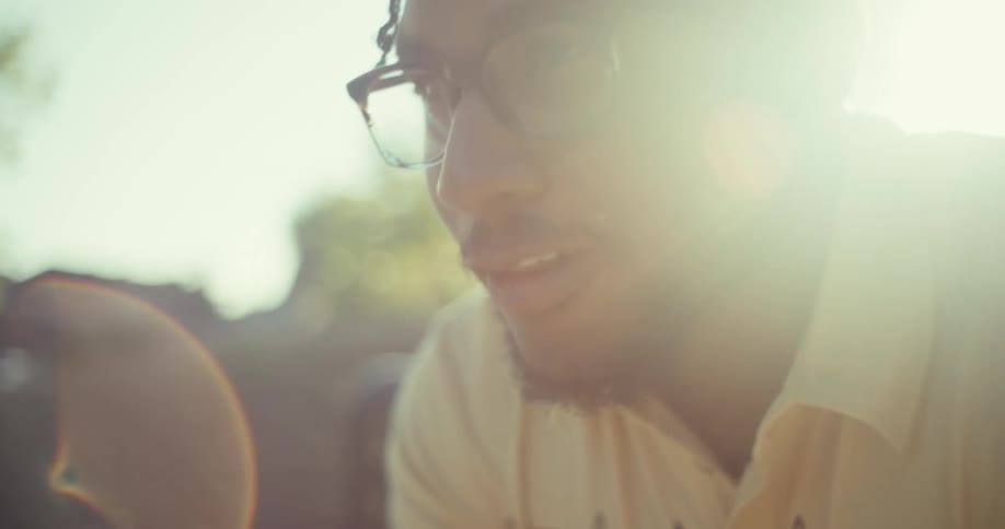 A person in glasses and a light shirt sits on a park bench under the sun