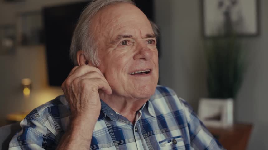An elderly man with glasses and an earbud smiles and speaks animatedly seemingly engaged in a video call or listening to something