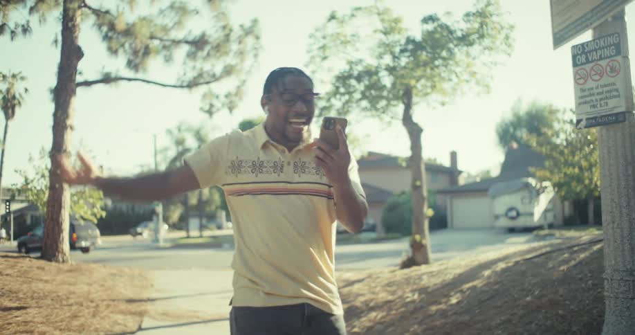 A man with braided hair glasses and white wireless earbuds walks outdoors on a sunny day he holds a smartphone and appears to be dancing or moving rhythmically smiling and looking at the phone