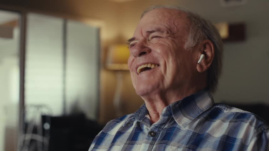Elderly man reacts happily on a phone call while sitting at a table in a living room during daytime