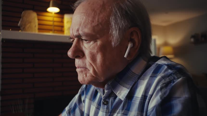 An elderly man sits indoors focused and working on his laptop