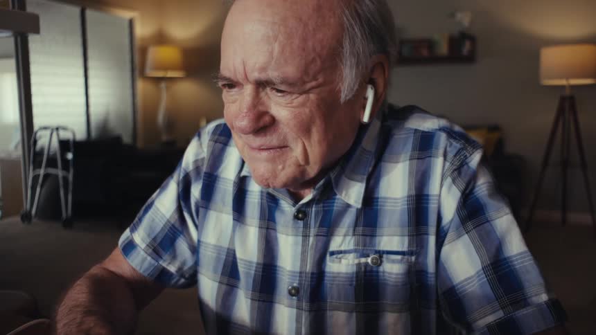 An elderly person in a plaid shirt and white earbuds sits indoors at a table with a laptop engaged in a phone call