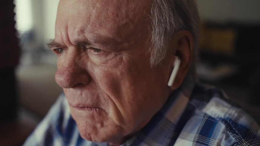 An elderly person in a plaid shirt and white earbuds sits indoors pausing midcall in visible shock after receiving bad news