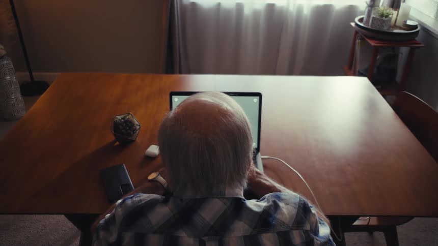 An elderly person sits at a wooden table using a laptop with a smartphone mouse and decorative object nearby