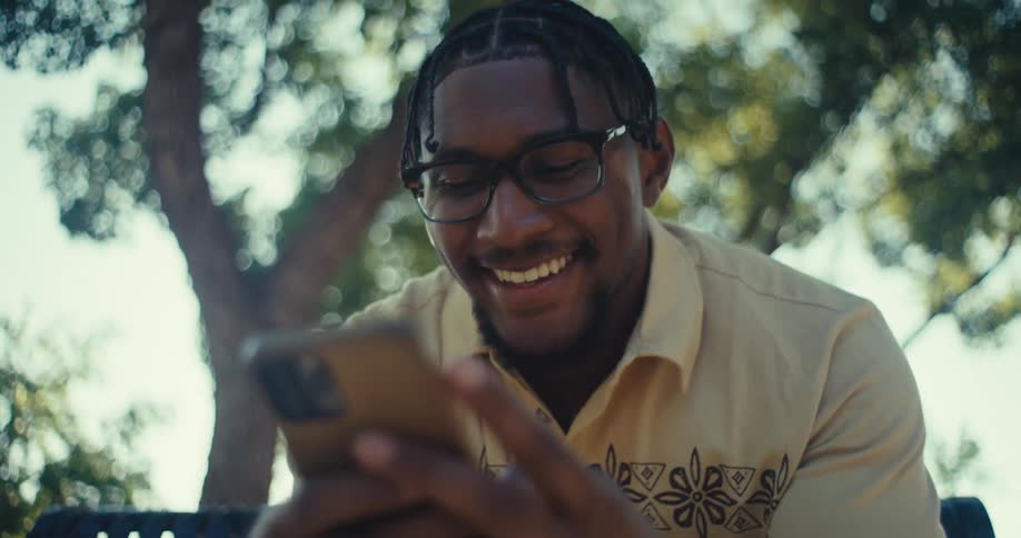 A person with braided hair and glasses wearing a lightcolored collared shirt is shown in a closeup they smile and nod appearing to be engaged in a conversation or reacting positively to something offcamera in an outdoor setting