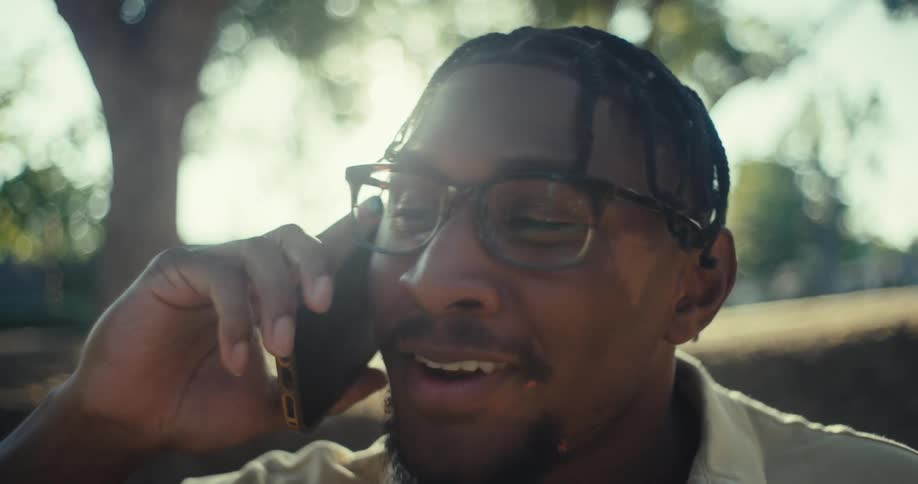 A smiling man sits on a park bench chatting on the phone beneath sunlit trees on a clear day