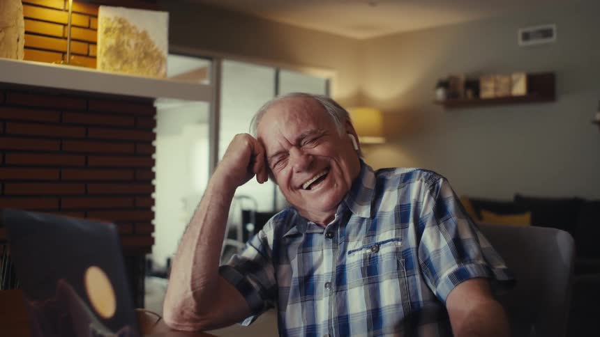 An elderly man wearing glasses and an earbud laughs heartily while resting his forehead with his hand seated at a table with a laptop