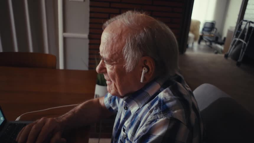An elderly man wearing earbuds is sitting in his living room at midday laughing while looking at his laptop monitor