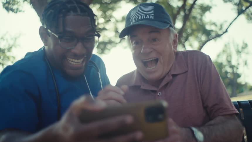 Nurse and an elderly man sitting on a bench under a tree celebrating happily watching on a smartphone