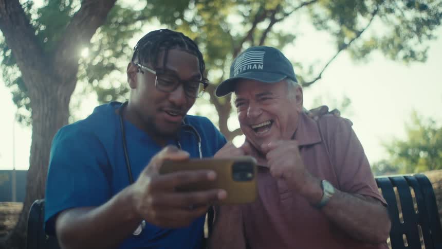A doctor wearing a blue robe with a stethoscope over his shoulder is sitting on a park bench beside an elderly man both are excitedly watching a game on the mobile device held by the doctor at midday