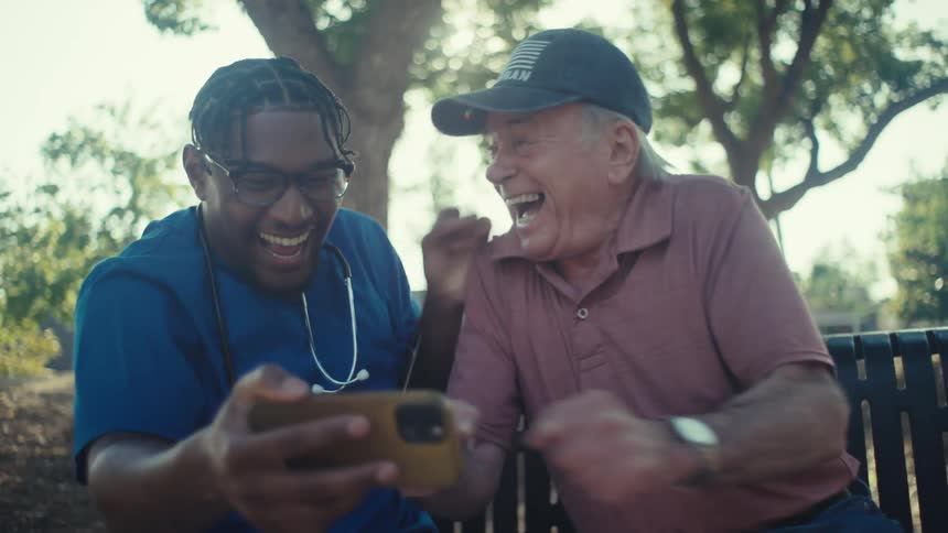 An elderly man wearing a veteran hat and a younger man possibly a caregiver sitting outdoors on a park bench they are both looking at a smartphone and smiling suggesting they are sharing a joyful moment or watching something amusing together