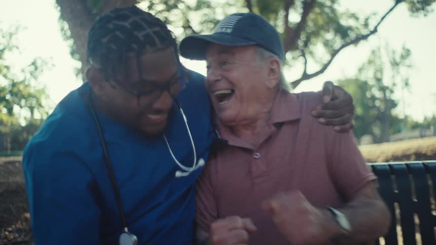 Young black male doctor celebrates with an elderly man after watching media content on a phone while sitting on a bench outdoors during daytime