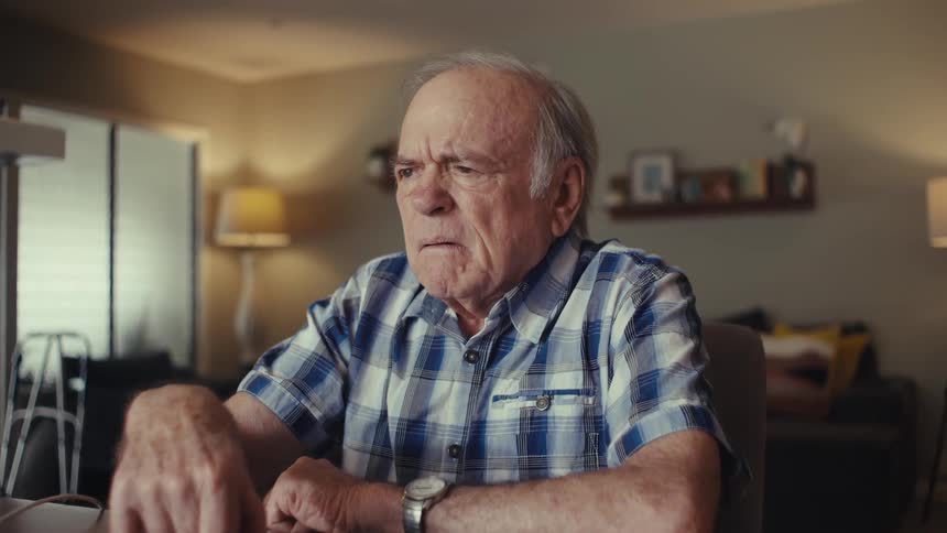 Elderly man reacts emotionally after taking a call while sitting at a table in a living room