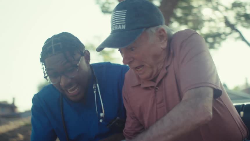 A doctor wearing a blue robe with a stethoscope over his shoulder is sitting on a bench in the park beside an elderly man both are filled with excitement as they watch a game on the mobile device held by the doctor at midday
