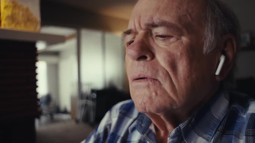 An elderly man is sitting in front of a laptop wearing earbuds and he looks concerned