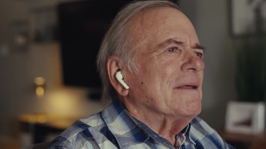 An elderly person in a plaid shirt and white earbuds sits indoors by a lamp engaged in a phone call