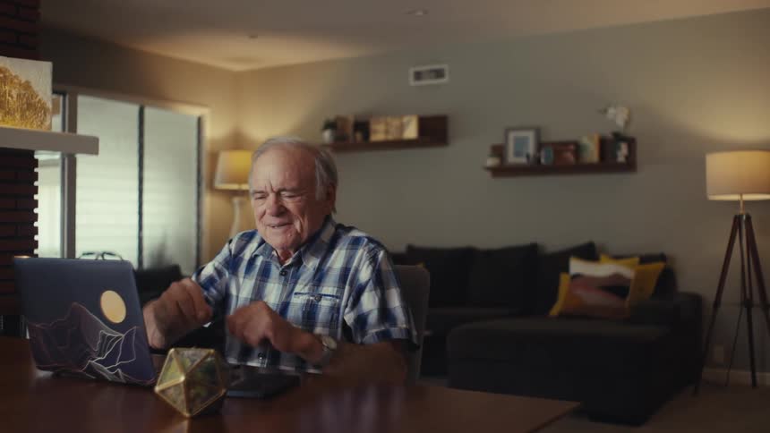 An elderly person in a plaid shirt and white earbuds sits indoors at a table with a laptop engaged in a phone call