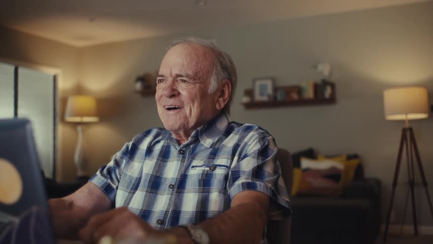 An elderly man wearing wireless earbuds laughs joyfully while video chatting on a laptop in a warmly lit living room
