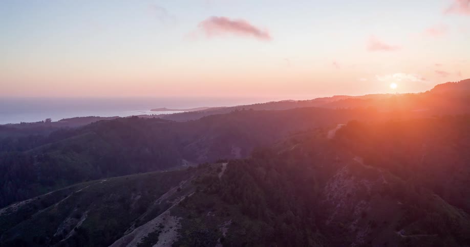 The sun sets over a range of forested hills that slope gently toward the ocean casting a warm orange glow across the landscape