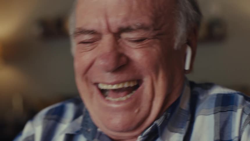 An elderly man with white hair wearing a blue and white plaid shirt and white wireless earbuds he transitions from a neutral expression to laughing heartily then returns to a more contemplative look