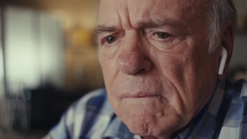 An elderly man wearing a plaid shirt and earbuds stares intently deep in thought