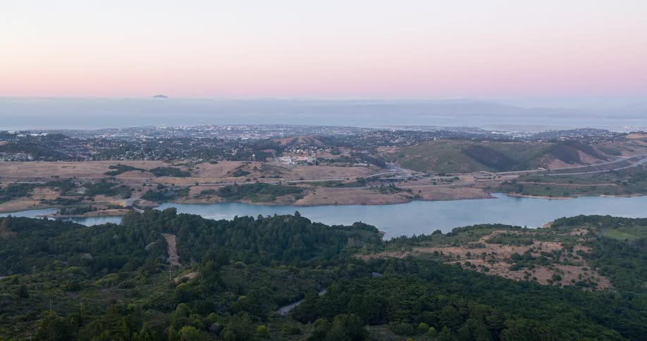 A wide aerial view of a hilly landscape with a river or reservoir forest and distant city at dawn