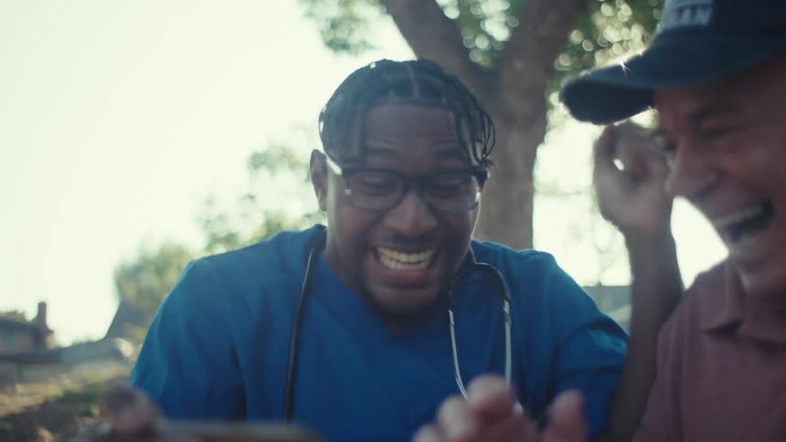 A healthcare professional in blue scrubs with a stethoscope sits beside an elderly man on a wooden bench both erupting with joy as they look at a phone