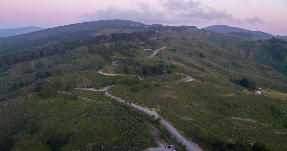 An aerial view captures winding roads cutting through rolling green hills and scattered trees under a dramatic cloudy sky