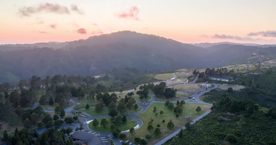 A winding road meanders through a lush hilly landscape at sunset with scattered trees and buildings dotting the scene