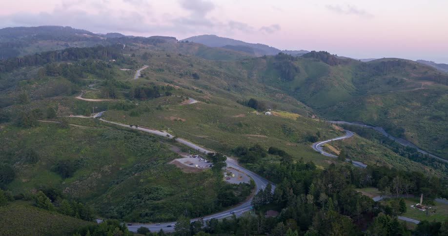 A winding road cuts through a range of green hills under a pastel dusk sky