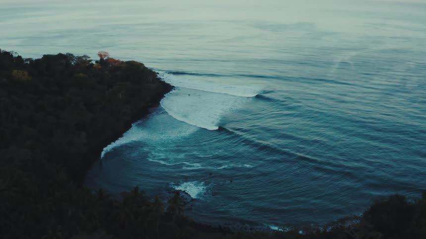 An aerial view captures the dramatic meeting of lush green cliffs and the vast serene ocean gentle waves can be seen rolling towards the shore with the light catching on the waters surface indicating either dawn or dusk