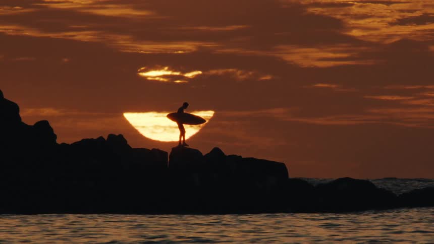 A surfer stands on rocks silhouetted against a dramatic sunset sky