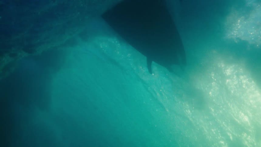 A surfboard glides overhead as sunlight filters through a curling underwater wave illuminating the swirling currents below
