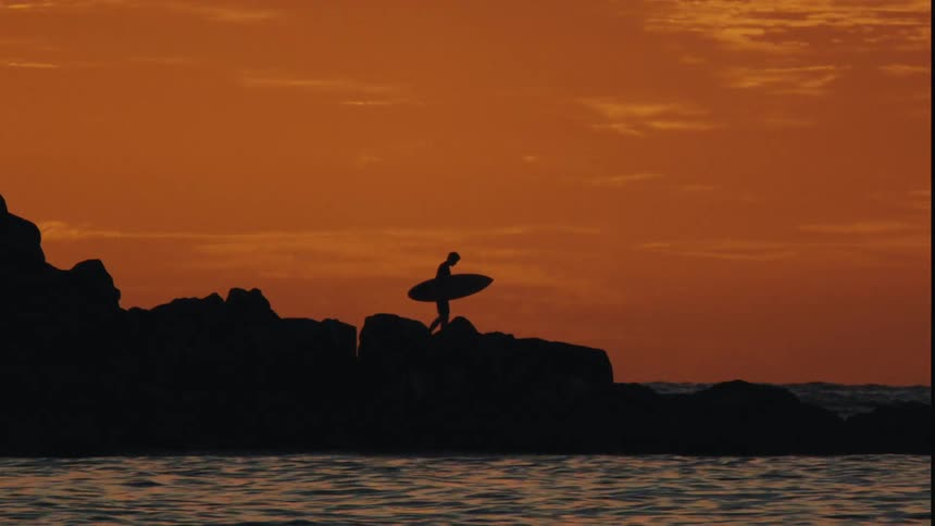 Surfer holding surfboard walking on rocks along the coast in afterglow of sunset