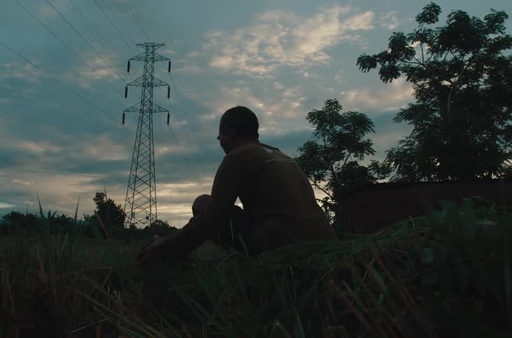 A man sits alone in a grassy field at dusk chopping grass silhouetted against a cloudy sky with power lines and a tall transmission tower in the background