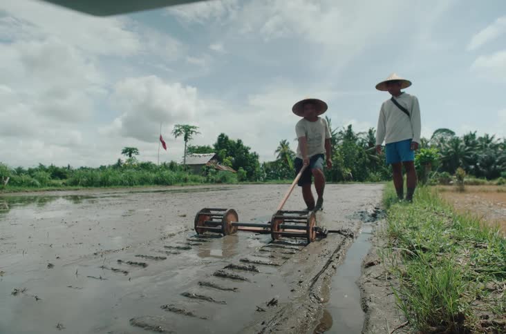 Two individuals in straw hats stand in a muddy field with one using a wooden tool to carve neat furrows in the soil while the other observes