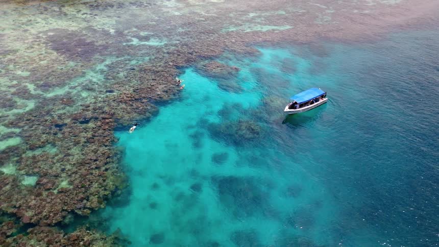 A boat is on the ocean water from an aerial view with the ocean floor and coral reefs visible at midday