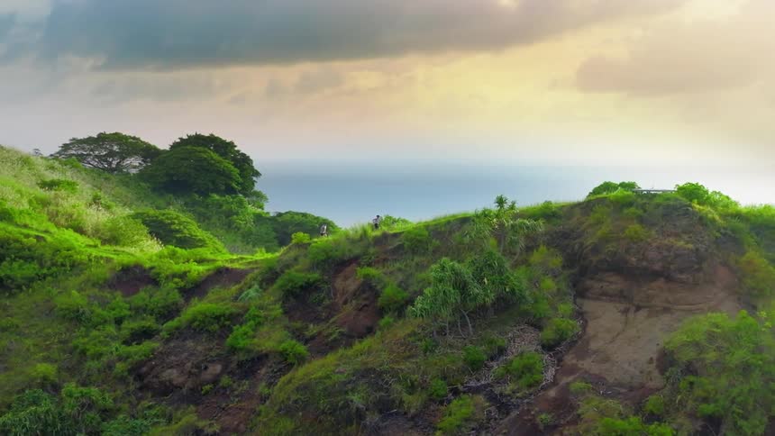 An aerial shot reveals two individuals walking along the ridge of a steep green cliff overlooking the ocean the sky is a mix of pale yellow and grey suggesting either early morning or late afternoon