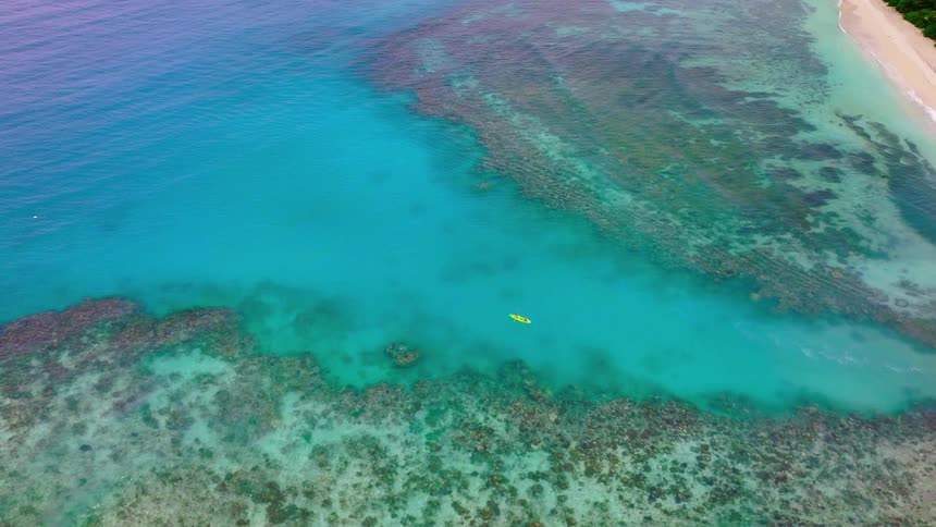 An aerial view captures a yellow kayak floating in a clear turquoise channel surrounded by dark coral reefs