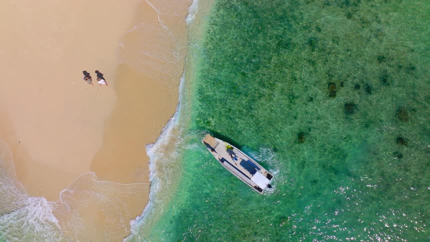 Aerial view of two people walking on the sand by a greenish sea with a yacht parked nearby
