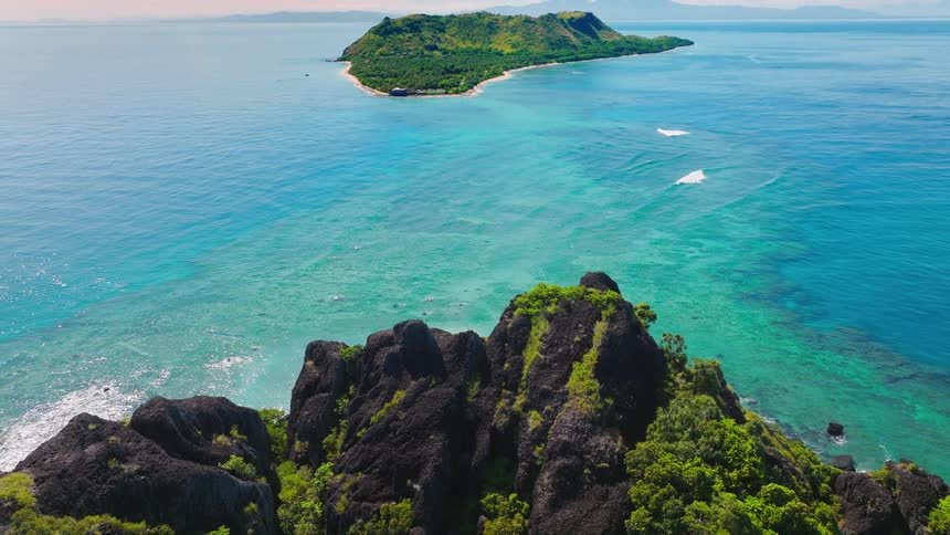 An aerial view showcases a vibrant tropical landscape featuring jagged dark green rocks covered in lush vegetation surrounded by strikingly clear turquoise ocean in the distance a small island with a white sandy beach is visible amidst the blue waters