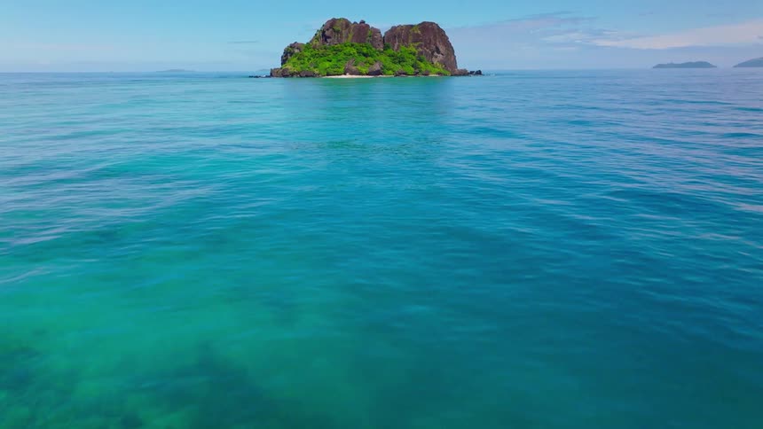 An aerial view shows a lush green island with a small white beach surrounded by clear blue ocean under a bright sky