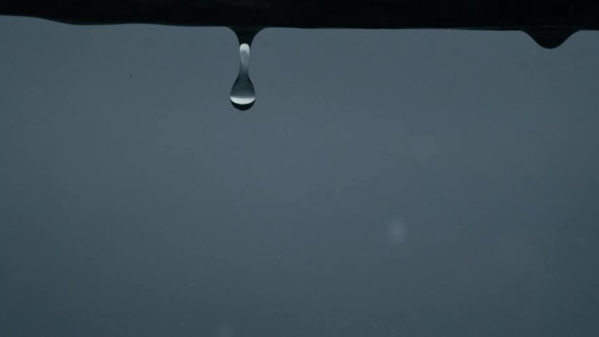 A water droplet falls from a dark horizontal edge as rain continues to fall