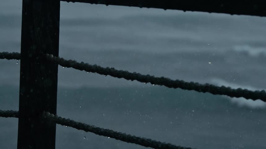 A wet textured rope likely part of a railing or fence has water droplets visible on its surface in the background is a dark body of water possibly the ocean due to rain