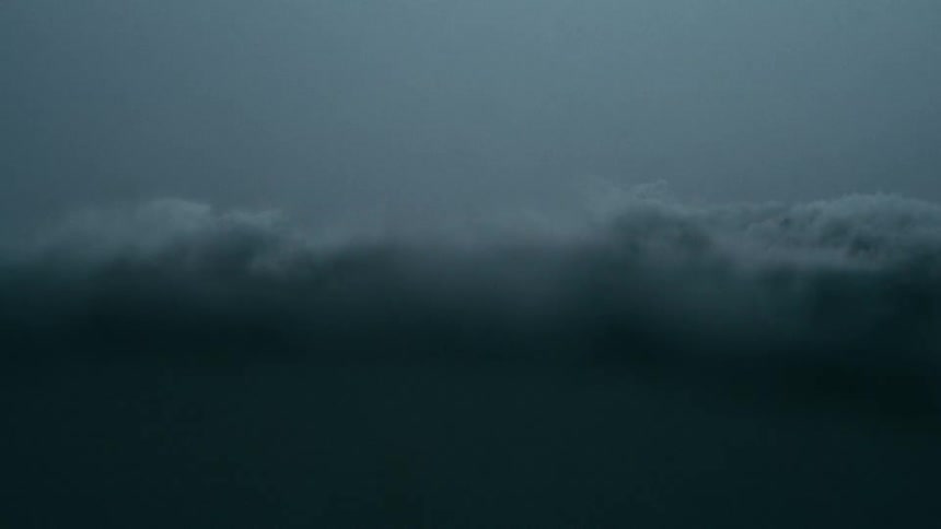Raindrops fall upon the surface of a large ocean wave under a gray sky